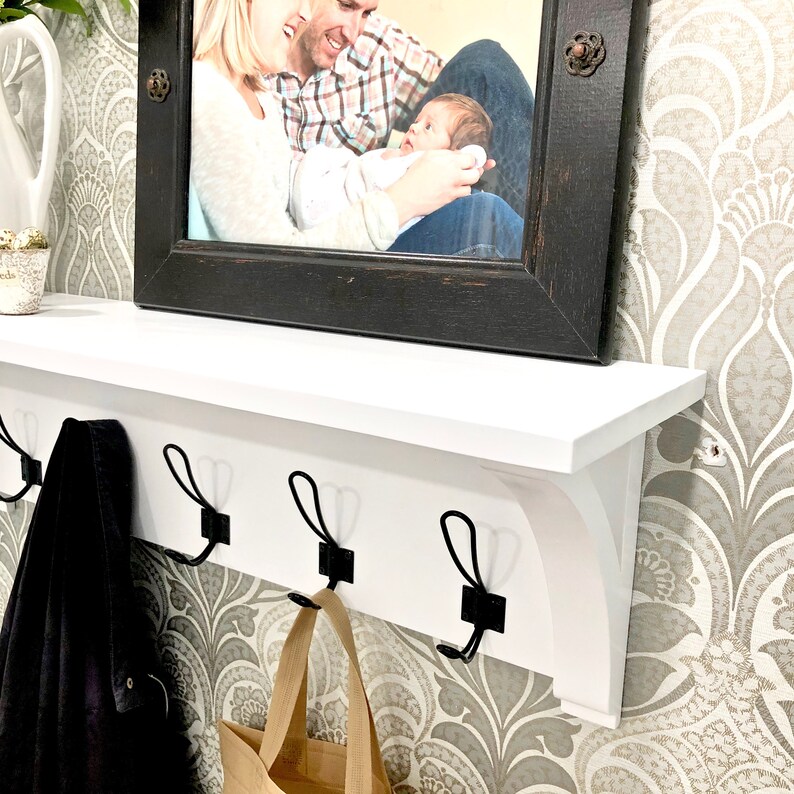 May include: A white wall-mounted shelf featuring three black metal hooks for hanging items. The shelf has a flat top surface and decorative brackets on the sides. A black wooden picture frame containing a photograph of a family is placed on top.
