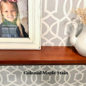 May include: A close-up of a wooden shelf stained with "Colonial Maple Stain". The shelf is decorated with a framed photo of a young girl and a white teapot with dried flowers.