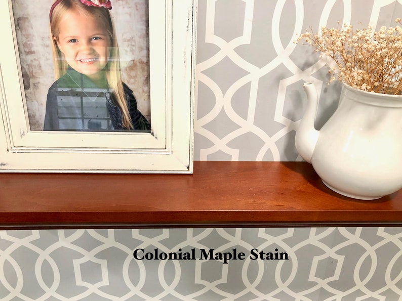 May include: A close-up of a wooden shelf stained with "Colonial Maple Stain". The shelf is decorated with a framed photo of a young girl and a white teapot with dried flowers.