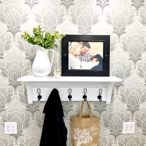May include: A white wall-mounted shelf with five black metal hooks, featuring a white ceramic pitcher filled with green foliage and a black rectangular picture frame containing a photograph of a family, with a dark coat and a tan tote bag hanging below.
