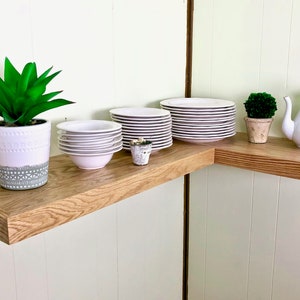 Puede incluir: Un juego de platos y cuencos blancos est&aacute;n apilados en un estante de esquina de madera marr&oacute;n claro. Una tetera blanca y una peque&ntilde;a planta en maceta est&aacute;n en el estante. Una planta en maceta m&aacute;s grande est&aacute; en el estante inferior.