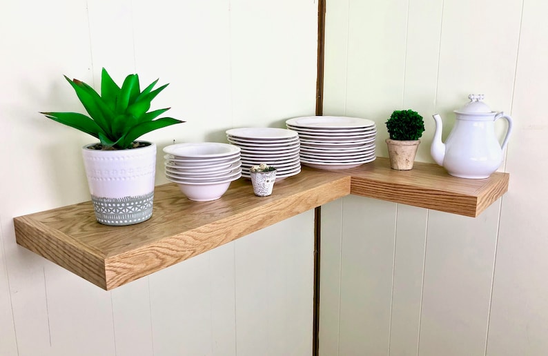 Puede incluir: Un juego de platos y cuencos blancos est&aacute;n apilados en un estante de esquina de madera marr&oacute;n claro. Una tetera blanca y una peque&ntilde;a planta en maceta est&aacute;n en el estante. Una planta en maceta m&aacute;s grande est&aacute; en el estante inferior.