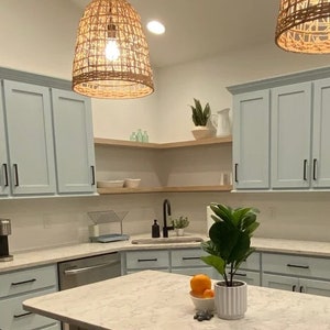 Puede incluir: Una cocina con armarios de color azul claro, una encimera blanca y un fregadero de acero inoxidable. Hay dos l&aacute;mparas colgantes de mimbre colgando del techo. Una planta en maceta se encuentra en la encimera.