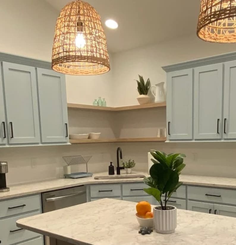 Puede incluir: Una cocina con armarios de color azul claro, una encimera blanca y un fregadero de acero inoxidable. Hay dos l&aacute;mparas colgantes de mimbre colgando del techo. Una planta en maceta se encuentra en la encimera.