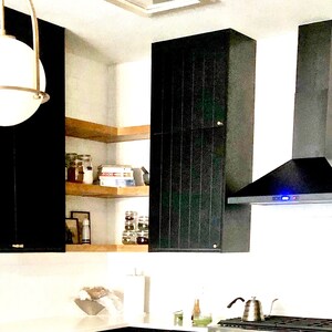 Puede incluir: Una campana extractora negra con una luz azul est&aacute; instalada sobre una cocina negra y de madera con estantes abiertos. La cocina tiene una encimera blanca y un fregadero de acero inoxidable.