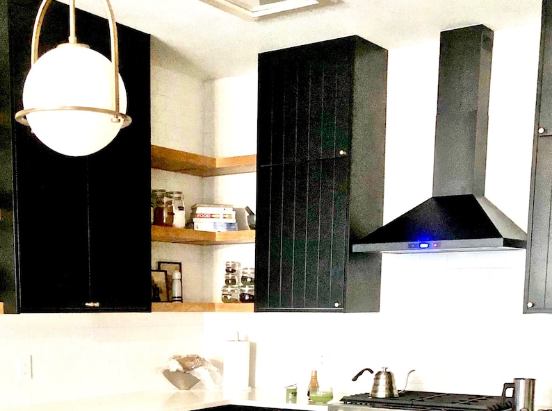 Puede incluir: Una campana extractora negra con una luz azul est&aacute; instalada sobre una cocina negra y de madera con estantes abiertos. La cocina tiene una encimera blanca y un fregadero de acero inoxidable.