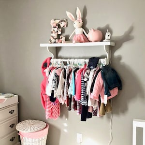 Puede incluir: Una estanter&iacute;a de madera blanca con un perchero de metal blanco adjunto. La estanter&iacute;a est&aacute; decorada con una figurita de cerdo rosa, un mu&ntilde;eco de conejo blanco y una letra E floral. El perchero est&aacute; lleno de ropa de beb&eacute; de varios colores.