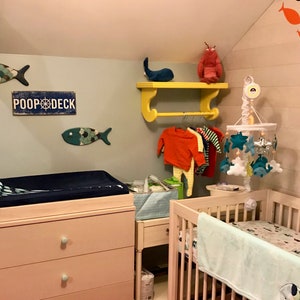 Puede incluir: Una habitaci&oacute;n de beb&eacute; con una c&oacute;moda rosa, una cuna blanca y un estante amarillo con un juguete de ballena y un juguete de langosta. El estante tiene un perchero con ropa de beb&eacute; colgada. La pared tiene un letrero que dice "Poop Deck" y un recorte de pescado. Hay un m&oacute;vil colgado sobre la cuna con estrellas, nubes y peces.