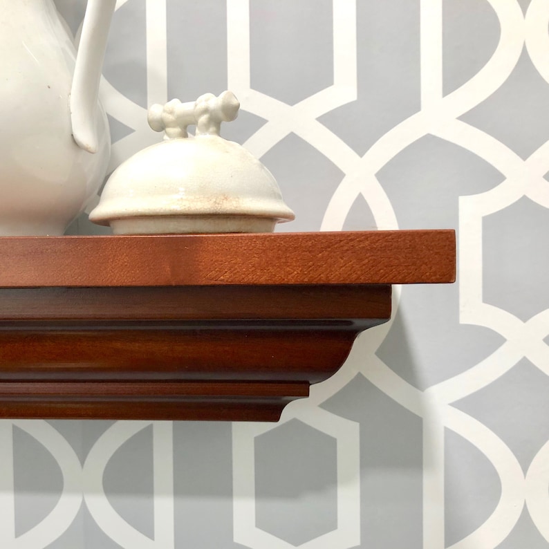 May include: A wooden shelf with a white ceramic lidded container and a white ceramic pitcher on top. The shelf has a decorative edge and is against a gray wall with a white geometric pattern.