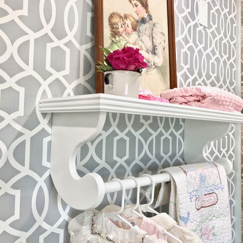 Puede incluir: Estante blanco con una barra blanca unida a la parte inferior. La estanter&iacute;a tiene un dise&ntilde;o decorativo de desplazamiento en los lados. La estanter&iacute;a est&aacute; montada en una pared con un papel tapiz de patr&oacute;n geom&eacute;trico gris y blanco. Hay ropa colgada en la barra y una manta rosa en la estanter&iacute;a.