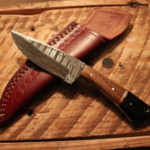 May include: A handmade knife with a patterned steel blade and a wooden handle with a black tip. The knife is resting on a wooden surface with a brown leather sheath.