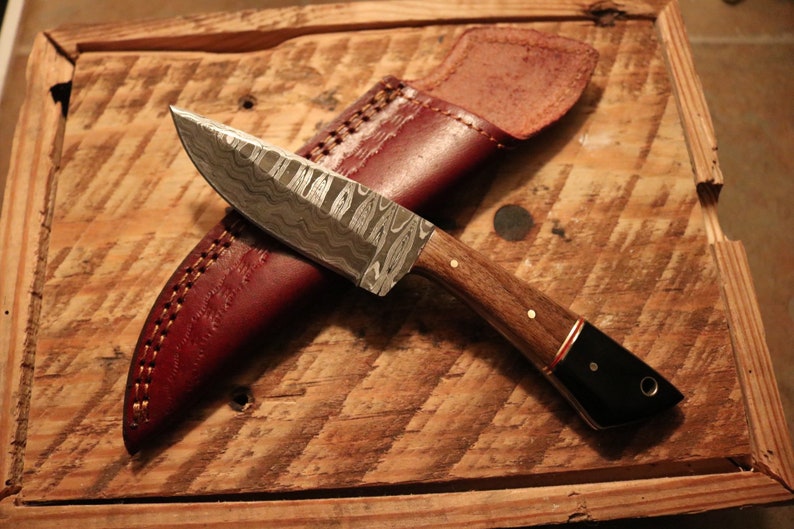 May include: A handmade knife with a patterned steel blade and a wooden handle with a black tip. The knife is resting on a wooden surface with a brown leather sheath.