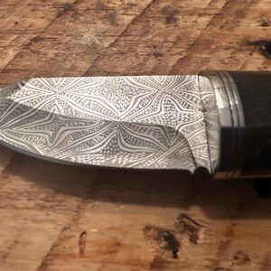 May include: A close-up of a knife with a patterned blade and a black handle. The blade is made of a metal with a silver finish and has a geometric pattern.