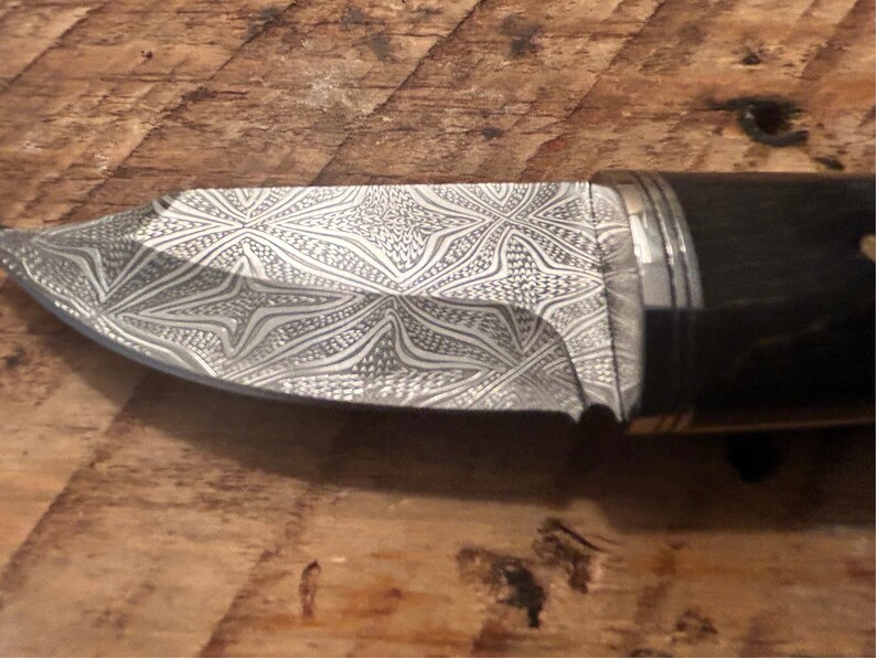May include: A close-up of a knife with a patterned blade and a black handle. The blade is made of a metal with a silver finish and has a geometric pattern.