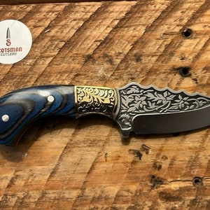 May include: A blue and black wood handle knife with a gold-colored floral design on the blade. The blade is etched with a floral pattern and has a sharp point. The knife is made by Scotsman Cutlery.