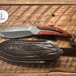 May include: A handcrafted knife with a Damascus steel blade and a wooden handle. The knife comes with a brown leather sheath with a feather design.