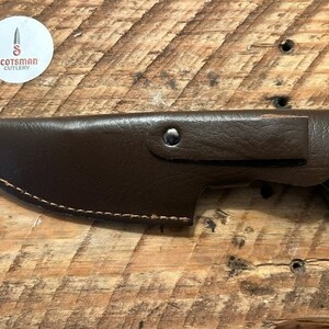 May include: A brown leather sheath with a stitched edge and a silver rivet. The sheath is holding a blue and black handled knife with a wood grain pattern.