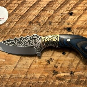 May include: A small, intricately designed knife with a blue and black wood handle. The blade features a floral pattern and a gold accent. The knife is made by Scotsman Cutlery.