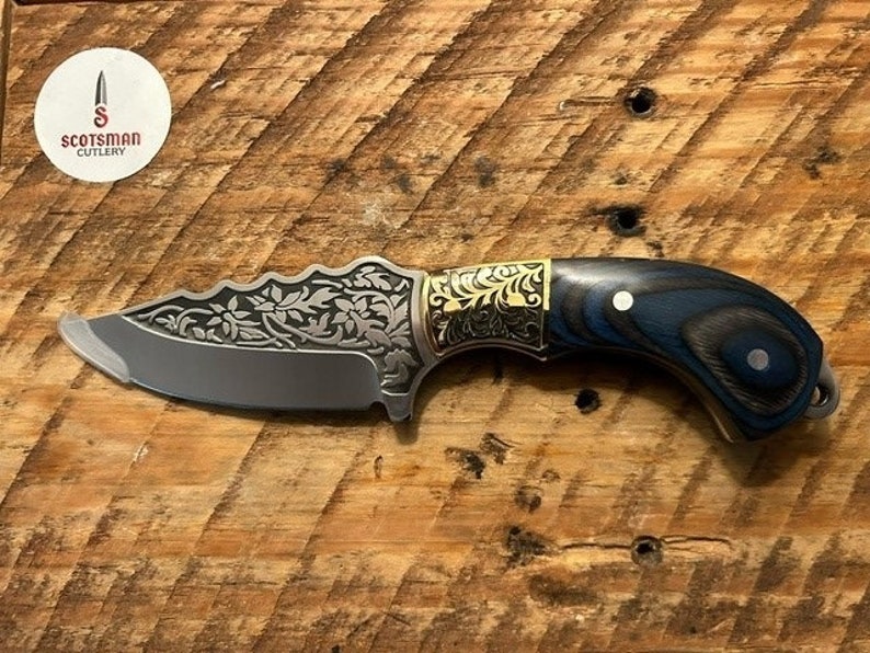 May include: A small, intricately designed knife with a blue and black wood handle. The blade features a floral pattern and a gold accent. The knife is made by Scotsman Cutlery.