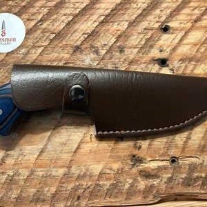 May include: A brown leather knife sheath with a blue and black wooden handle knife. The sheath has a stitched edge and a snap closure. The knife handle has a blue and black wood grain pattern.