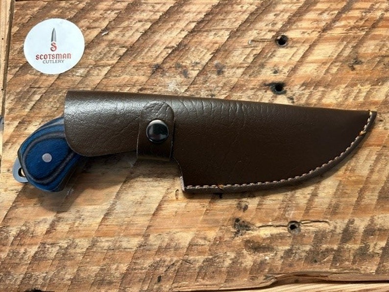 May include: A brown leather knife sheath with a blue and black wooden handle knife. The sheath has a stitched edge and a snap closure. The knife handle has a blue and black wood grain pattern.