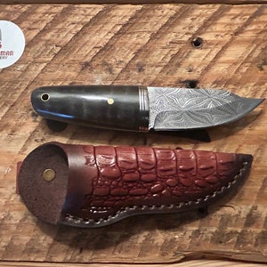May include: A small, black-handled knife with a patterned blade and a brown leather sheath. The knife is made by Scotsman Cutlery.