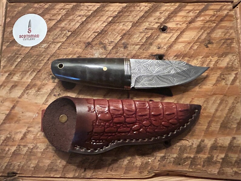 May include: A small, black-handled knife with a patterned blade and a brown leather sheath. The knife is made by Scotsman Cutlery.