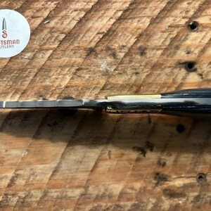 May include: A black and gold pocket knife with a wood handle. The knife has a serrated blade and a gold accent on the handle. The knife is made by Scotsman Cutlery.