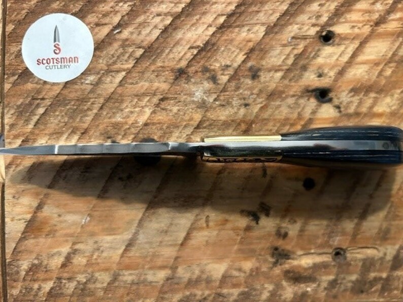 May include: A black and gold pocket knife with a wood handle. The knife has a serrated blade and a gold accent on the handle. The knife is made by Scotsman Cutlery.