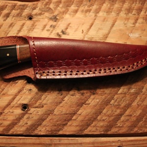 May include: A brown leather knife sheath with a decorative stitching pattern. The sheath is closed and a knife is inside.
