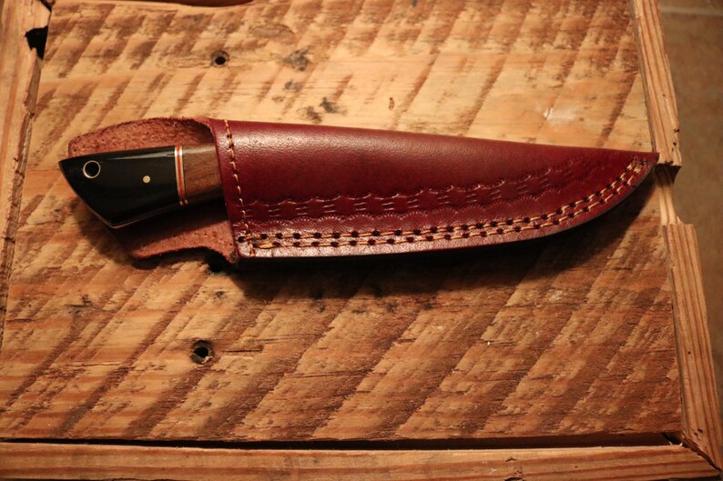 May include: A brown leather knife sheath with a decorative stitching pattern. The sheath is closed and a knife is inside.