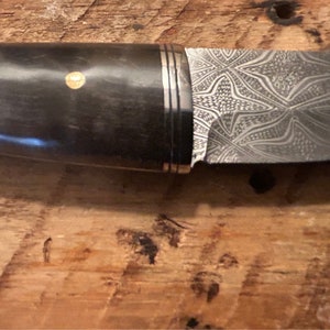 May include: A close-up of a knife with a black handle and a patterned steel blade. The handle has a gold accent.