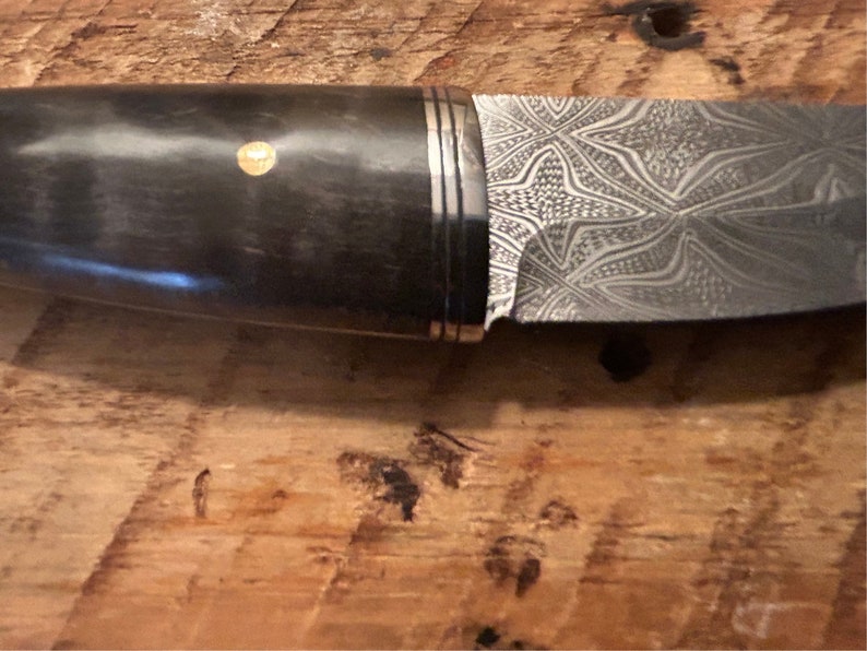 May include: A close-up of a knife with a black handle and a patterned steel blade. The handle has a gold accent.
