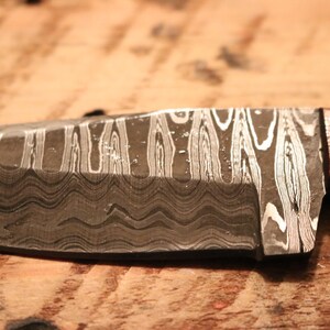 May include: A close-up of a knife with a wavy, patterned blade and a wooden handle. The blade is made of a metal with a silver and black pattern.