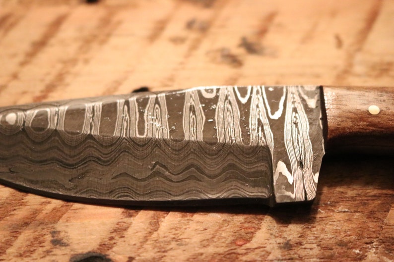 May include: A close-up of a knife with a wavy, patterned blade and a wooden handle. The blade is made of a metal with a silver and black pattern.