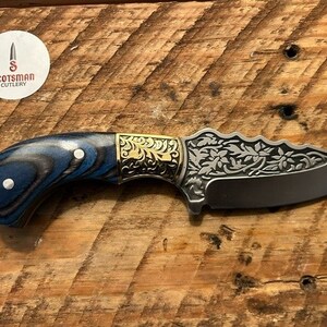 May include: A blue and black wood handle knife with a gold floral design on the blade. The blade is etched with a floral pattern and has a sharp point. The knife is made by Scotsman Cutlery.