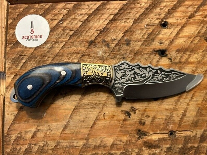 May include: A blue and black wood handle knife with a gold floral design on the blade. The blade is etched with a floral pattern and has a sharp point. The knife is made by Scotsman Cutlery.