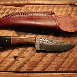 May include: A small, handmade knife with a patterned steel blade and a wooden handle. The knife is in a brown leather sheath.