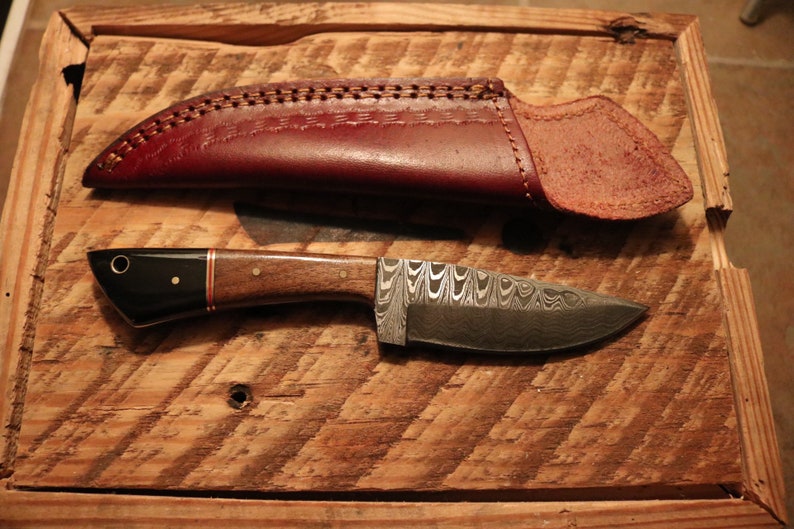 May include: A small, handmade knife with a patterned steel blade and a wooden handle. The knife is in a brown leather sheath.