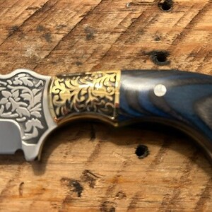 May include: A close-up of a knife with a blue and black wood handle, gold accents, and a silver blade with an engraved floral design.