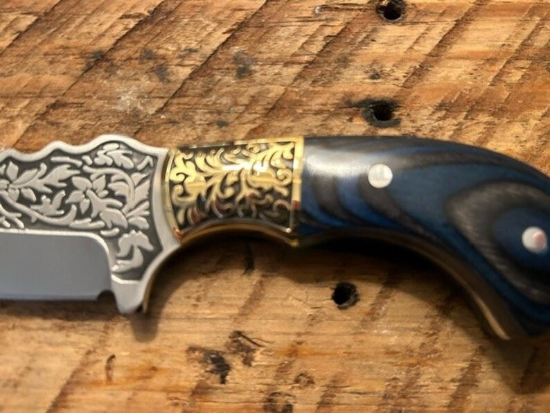May include: A close-up of a knife with a blue and black wood handle, gold accents, and a silver blade with an engraved floral design.
