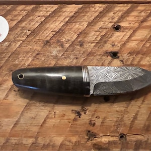 May include: A black and silver knife with a patterned blade and a black handle with gold accents. The knife is on a wooden surface.
