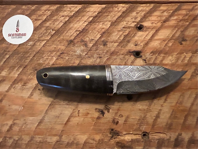 May include: A black and silver knife with a patterned blade and a black handle with gold accents. The knife is on a wooden surface.