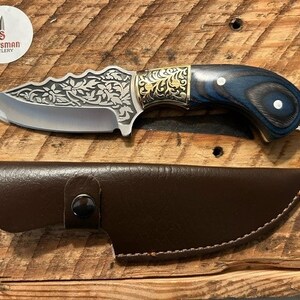 May include: A small, silver knife with a decorative floral pattern on the blade. The handle is made of dark wood with blue accents and gold accents. The knife comes with a brown leather sheath.