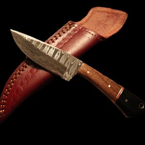 May include: A small, handcrafted knife with a patterned steel blade and a wooden handle. The knife is in a brown leather sheath.