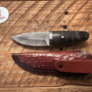May include: A small, silver knife with a patterned blade and a dark wood handle. The knife is in a brown leather sheath with a crocodile pattern.