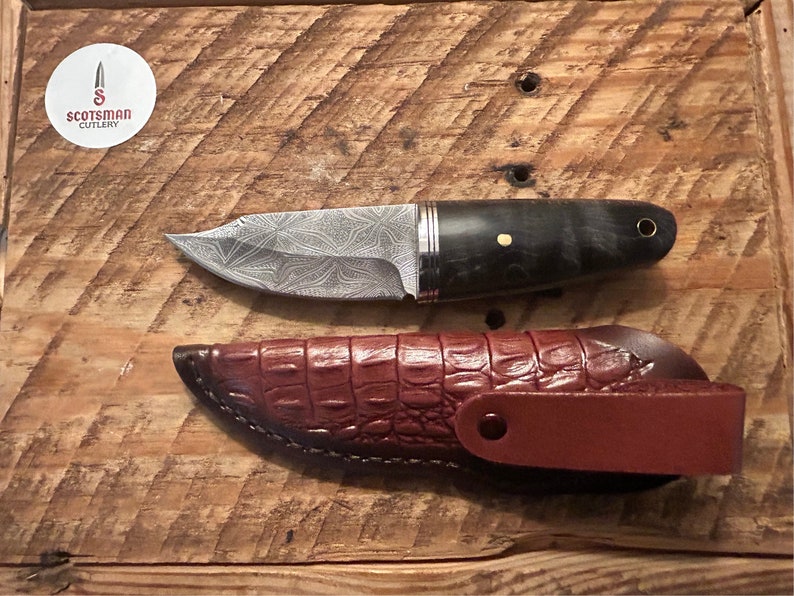 May include: A small, silver knife with a patterned blade and a dark wood handle. The knife is in a brown leather sheath with a crocodile pattern.