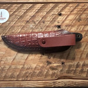 May include: A brown leather knife sheath with a crocodile pattern. The sheath has a brown leather strap with a metal snap closure. The knife is not visible in the image.