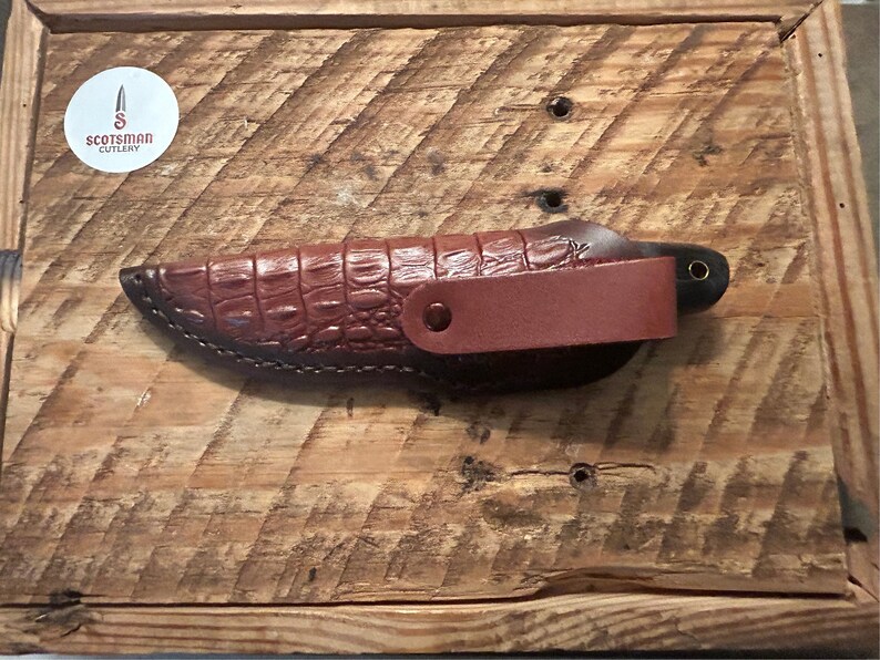 May include: A brown leather knife sheath with a crocodile pattern. The sheath has a brown leather strap with a metal snap closure. The knife is not visible in the image.