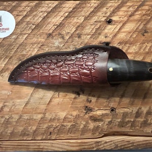 May include: A brown leather knife sheath with a crocodile pattern, a black and brown knife with a gold accent, and the text "Scotsman Cutlery" on a white circle.
